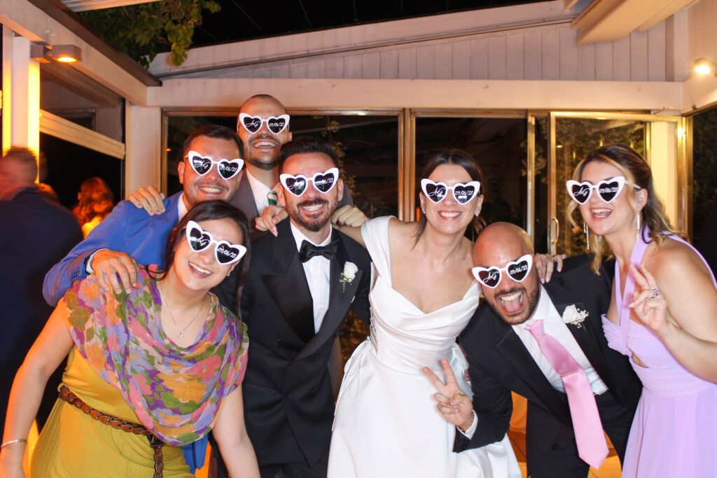 Sposi con occhiali da sole buffi insieme ad amici con gli stessi occhiali in posa davanti a un photo booth matrimonio
