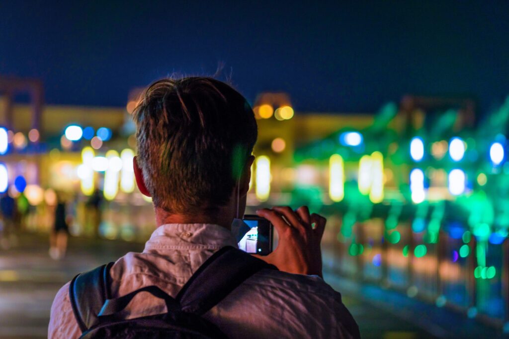 Uomo di spalle che scatta una foto notturna con lo smartphone in una città illuminata, tra luci colorate e sfondo bokeh, esempio di fotografia al buio con ricerca della luce.
